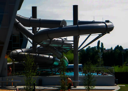 Eine graue Röhrenrutsche im Außenbereich eines Schwimmbades, mehrere Menschen sind im Schwimmbecken.