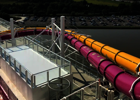 Wasserrutschen in Pink und Gelb führen vom Einstiegsturm eines Kreuzfahrtschiffes nach unten.