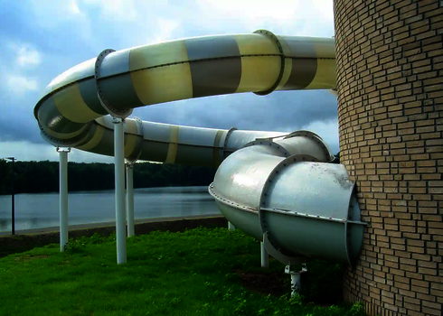 Gestreifte Wasserrutsche mit Fassadendurchdringung führt aus rundem Backsteingebäude am Wasser.