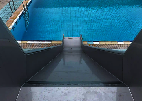 Die Kastenrutsche aus Edelstahl führt gerade in ein Schwimmbecken mit blauem Wasser.