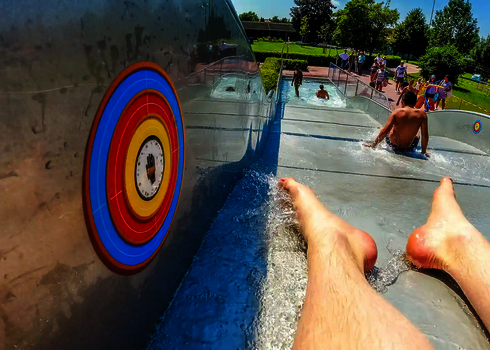 Bunter Interactivity Button, daneben Füße einer rutschenden Person und Freibad im Hintergrund.