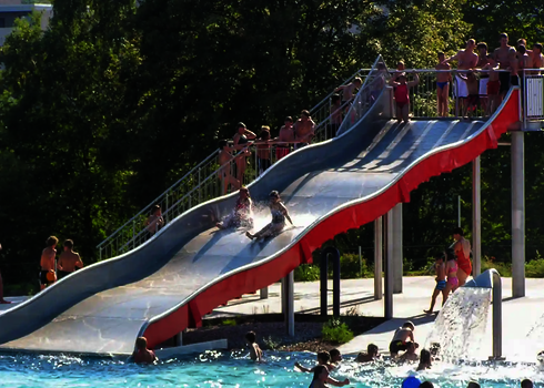 Viele Menschen in einem Schwimmbad, einige davon rutschen eine Breitwellenrutsche herunter.