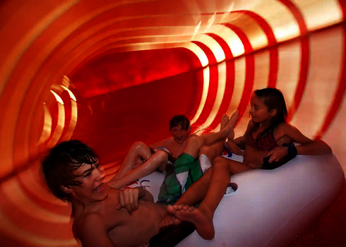 Eine orangene Wasserrutsche von innen, mit drei Personen auf einem weißen Rutschreifen.