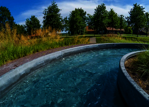 Kurve des Wildwasserflusses vor parkähnlicher Landschaft mit Bäumen und Häusern im Hintergrund.