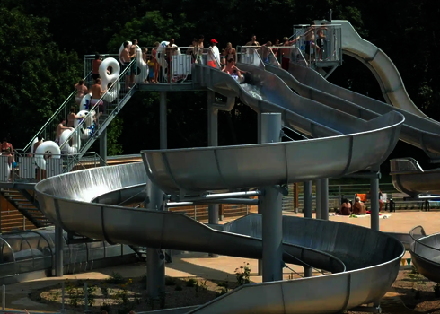 Unterschiedliche Wasserrutschen aus Edelstahl, Badegäste stehen mit und ohne Rutschreifen an.