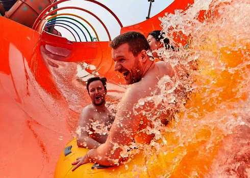 Vier Erwachsene fahren lachend durch die orangene Rutsche RR2700, Wasser spritzt hoch.