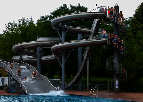 Mehrere Personen rutschen gleichzeitig eine Breitwellenrutsche herunter, daneben befindet sich ein Rutschenkomplex aus Edelstahl, bei dem mehrere Personen anstehen.