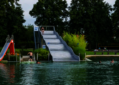 Eine Breitwellenrutsche und eine weitere Wasserrutsche in einer grün bewachsenen Umgebung, mit einigen Badegästen im Wasser. 