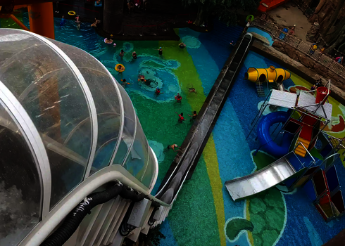 Blick von oben auf Rutsche und bunt gestalteten Wasserbereich mit spielenden Kindern.