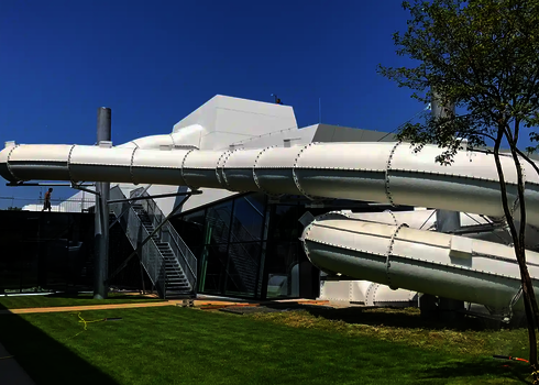 Weiße Wasserrutsche außerhalb eines Glasgebäudes, vor einer Wiese.