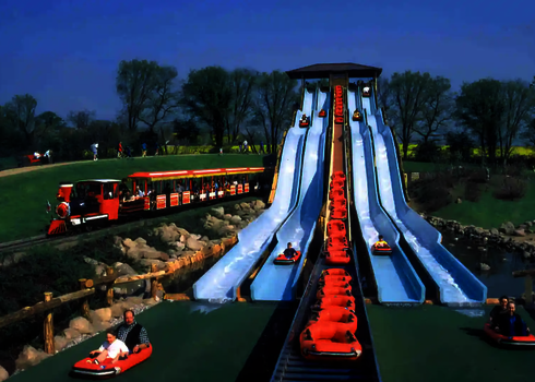 Freizeitpark unter blauem Himmel, vier parallel laufende Bahnen der Harakiri Wasserrutsche, dahinter eine kleine Eisenbahn.