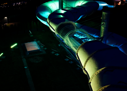 Außenaufnahme einer leuchtenden Röhrenrutsche mit transparentem Abschnitt bei Nacht.