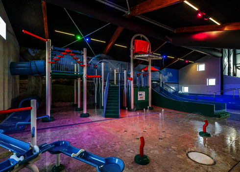 Kinderbereich in einem Hallenbad mit bunter Beleuchtung, Wasserrutschen in grün und blau und einem roten Wassereimer.