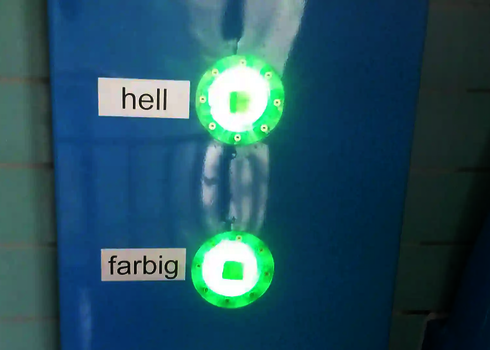 Zwei beleuchtete Tasten mit der Beschriftung „hell“ und „farbig“ an einer blauen Wand.