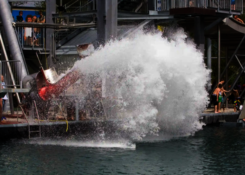Großer Wasserschwall vor einem Cannonball in einem Freibad, im Hintergrund sind Kinder an einer Kletterwand.