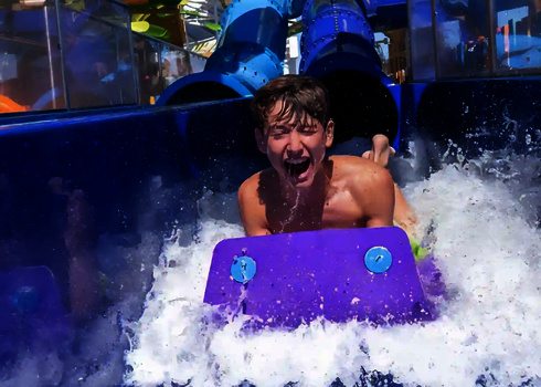 Boy slides down a water slide on a purple slide mat and lands in the water laughing.
