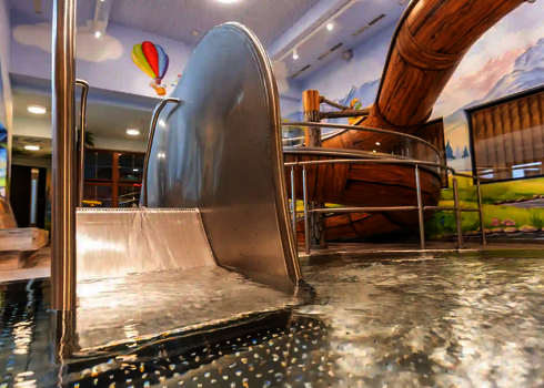  Edelstahlrutsche im Kinderbecken mit Wasserzufluss, dahinter eine große Rutsche in Holzoptik.