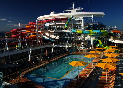 Kreuzfahrtschiffdeck mit Poolbereich, gelben Sonnenschirmen und bunten Wasserrutschen im Hintergrund.