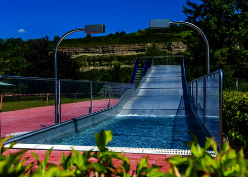 Breitwellenrutsche mit blauer Treppe und Auslaufbecken vor einem grün bewachsenen Berg.