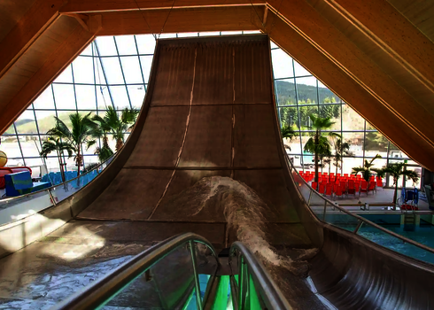 Blick auf die steile Rampe der Boomerang-Rutsche in einer hellen Schwimmhalle mit Palmen.