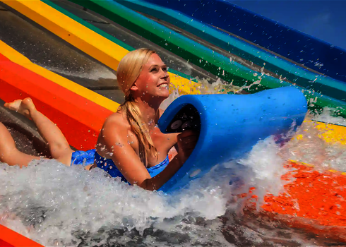 Young woman slides down a colourful matracer water slide with a blue mat, laughing as she hits the water.