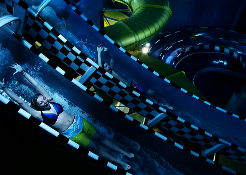 Zwei Personen rutschen nebeneinander durch transparente Röhren mit Zielflaggenmuster.