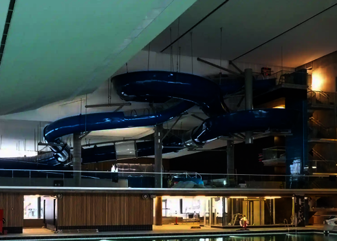 Schwimmbad mit einer blauen, geschwungenen Wasserrutsche, die von Stahlseilen getragen wird.