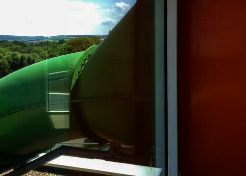 Grüne Wasserrutsche tritt durch Glasfassade neben orangefarbener Wand ins Freie.