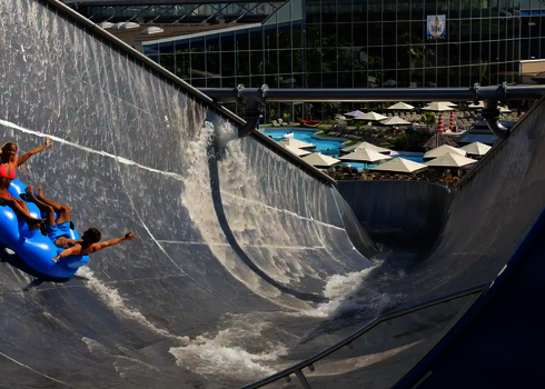 Menschen rutschen jubelnd eine Edelstahlwasserrutsche, im Hintergrund ein Gebäude mit Glasfassade.
