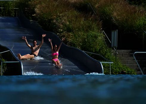 Zwei lachende Personen rutschen mit erhobenen Armen eine Breitwellenrutsche aus Edelstahl hinunter.