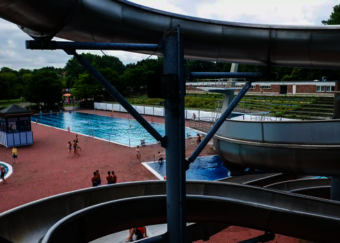 Blick auf ein Freibad mit Schwimmbecken und Badegästen, im Vordergrund eine Edelstahl-Wasserrutsche.