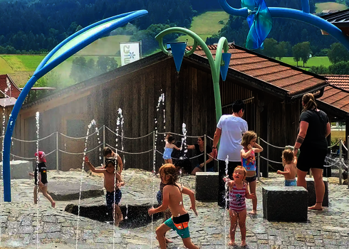 Kinder auf Wasserspielplatz mit Wie-Spray Elementen mit wäldlichem Hintergrund.