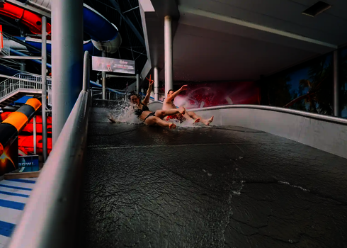 Zwei Personen rutschen mit erhobenen Händen eine Breitwellenrutsche in einem Wasserpark herunter.