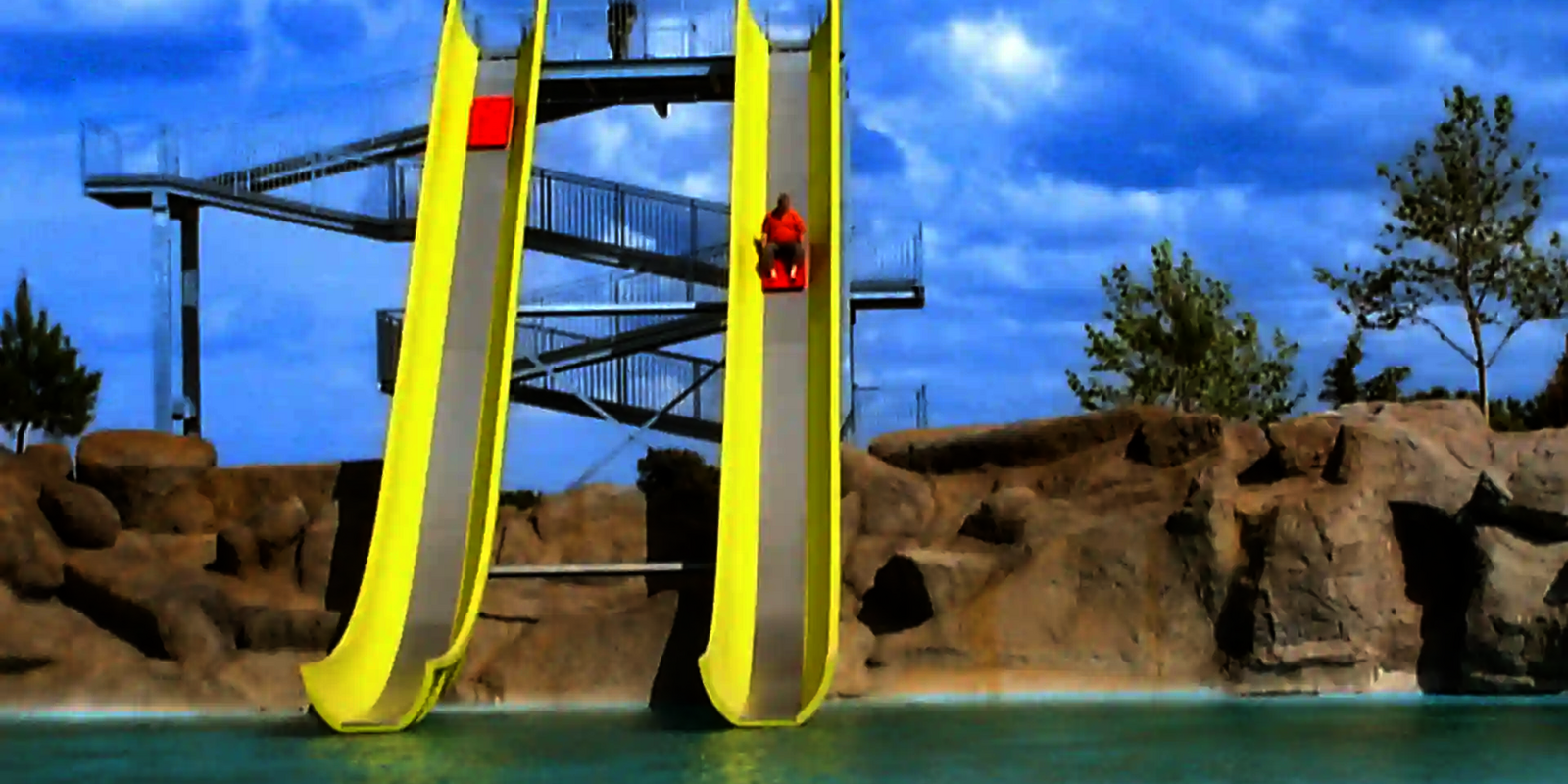 Eine Person rutscht auf einem Boot die gelbe Aqua Shuttle Wasserrutsche herunter, unter freiem, blauen Himmel.