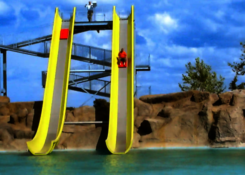 Eine Person rutscht auf einem Boot die gelbe Aqua Shuttle Wasserrutsche herunter, unter freiem, blauen Himmel.