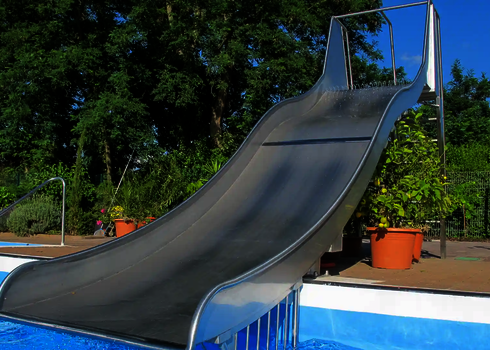 Eine Breitwellenrutsche in an einem Schwimmbecken vor grünen Bäumen und Pflanzenkübeln.
