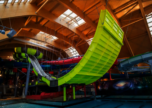 Eine gelbe Boomerang-Rutsche wird über Seile an der Hallendecke fixiert und endet über dem Wasser.