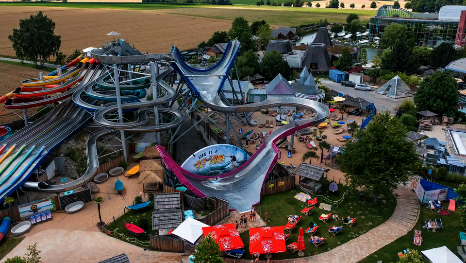 Luftaufnahme eines Wasserparks mit einem Sidewinder, einem Urknall und Liegewiese im Vordergrund.