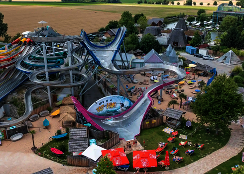 Luftaufnahme eines Wasserparks mit einem Sidewinder, einem Urknall und Liegewiese im Vordergrund.