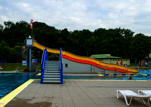 Sicht auf ein Schwimmbad mit einer gelben Breitwellenrutsche und einer blauen Treppe.
