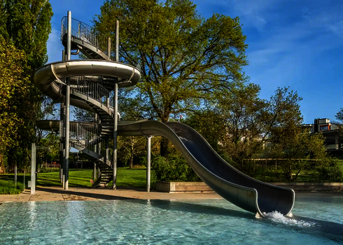 Eine Edelstahlrutsche mit Spiralaufgang endet in einem Becken, umgeben von Natur.