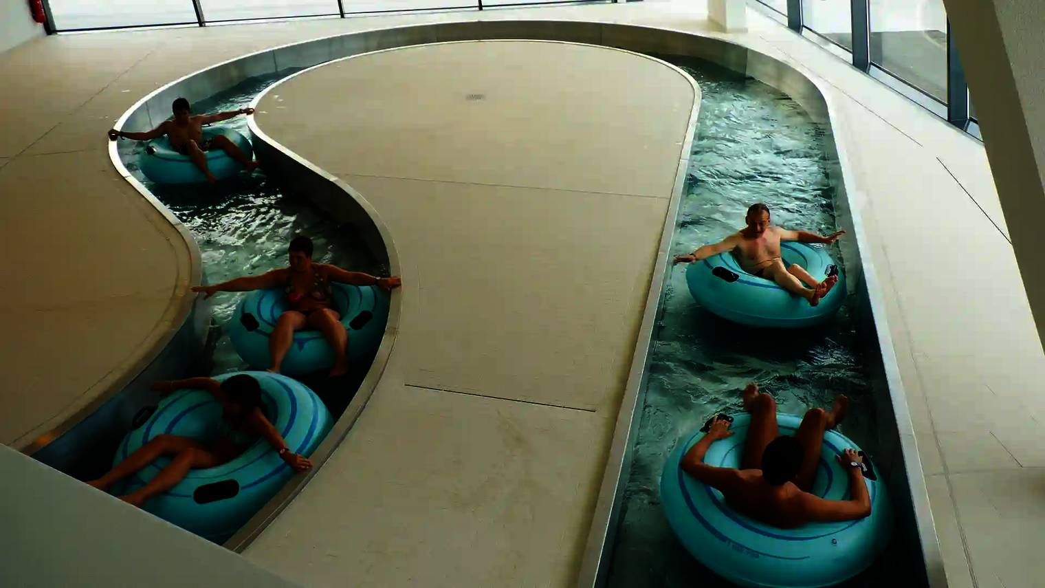 Mehrere Personen treiben auf Rutschreifen durch den Lazy River mit Panoramafenstern.