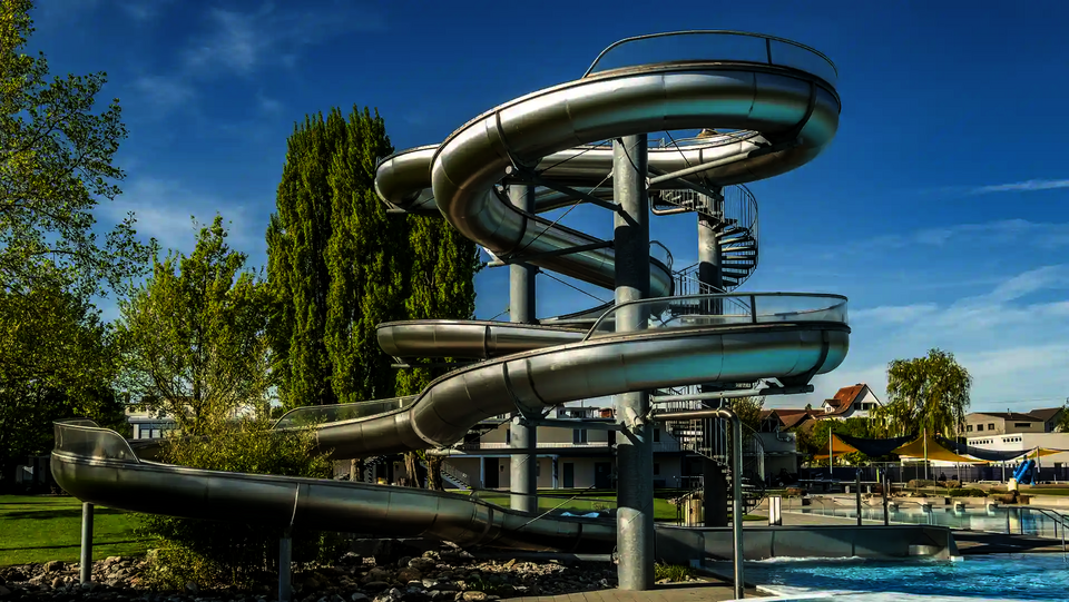 Mehrere Edelstahlrutschen verlaufen spiralförmig an einem Rutschenturm in einem Freibad.