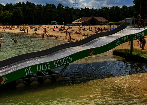 Seitenansicht einer grünen Breitwellenrutsche, die sich in einem Freibad mit Strandabschnitt befindet.