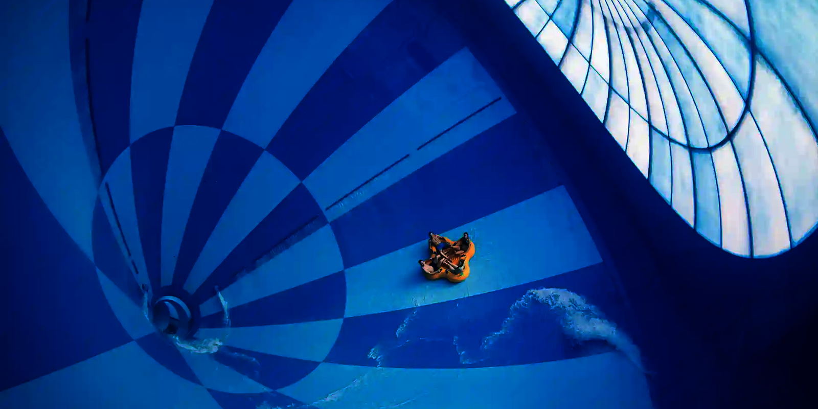 Große Wasserrutsche mit blauem und dunklem Muster, auf der ein Schlauchboot mit mehreren Personen fährt
