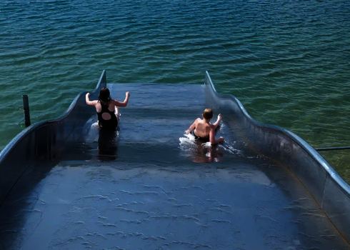 Blick von oben, wie zwei Personen eine Breitwellenrutsche in einem See herunterrutschen.