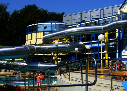 Eine große Edelstahlrutsche befindet sich im Außenbereich eines Schwimmbades, ein Kind mit Schwimmflügeln steht daneben.