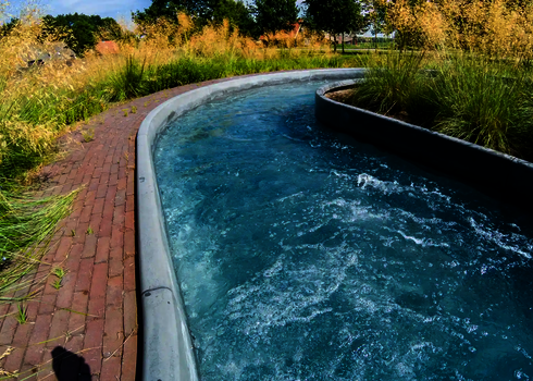 Sanft geschwungene Kurve des Wildwasserflusses neben gepflastertem Weg mit hohen Gräsern.