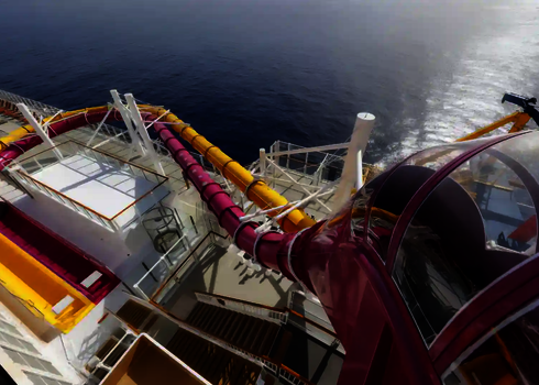 Blick aus der Vogelperspektive auf den Startbereich und den weiteren Rutschverlauf einer pinken und gelben Wasserrutsche auf einem Kreuzfahrtschiff.