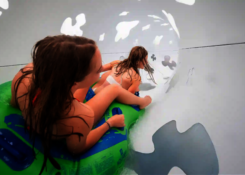 Innenansicht einer Wasserrutsche in der zwei Frauen in einem grünen Reifen rutschen.